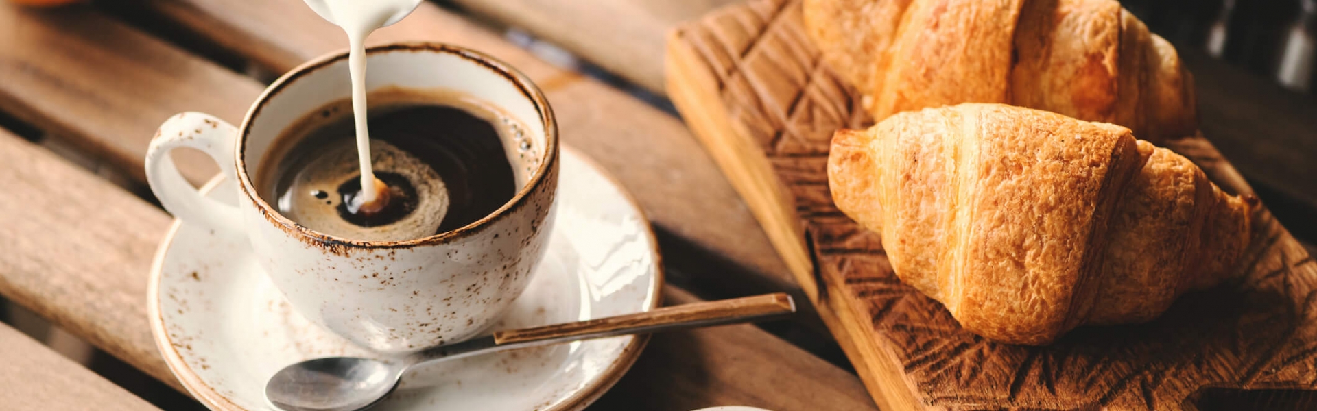Pouring cream into cup of black coffee. Tasty breakfast table set with croissants, jam and coffee